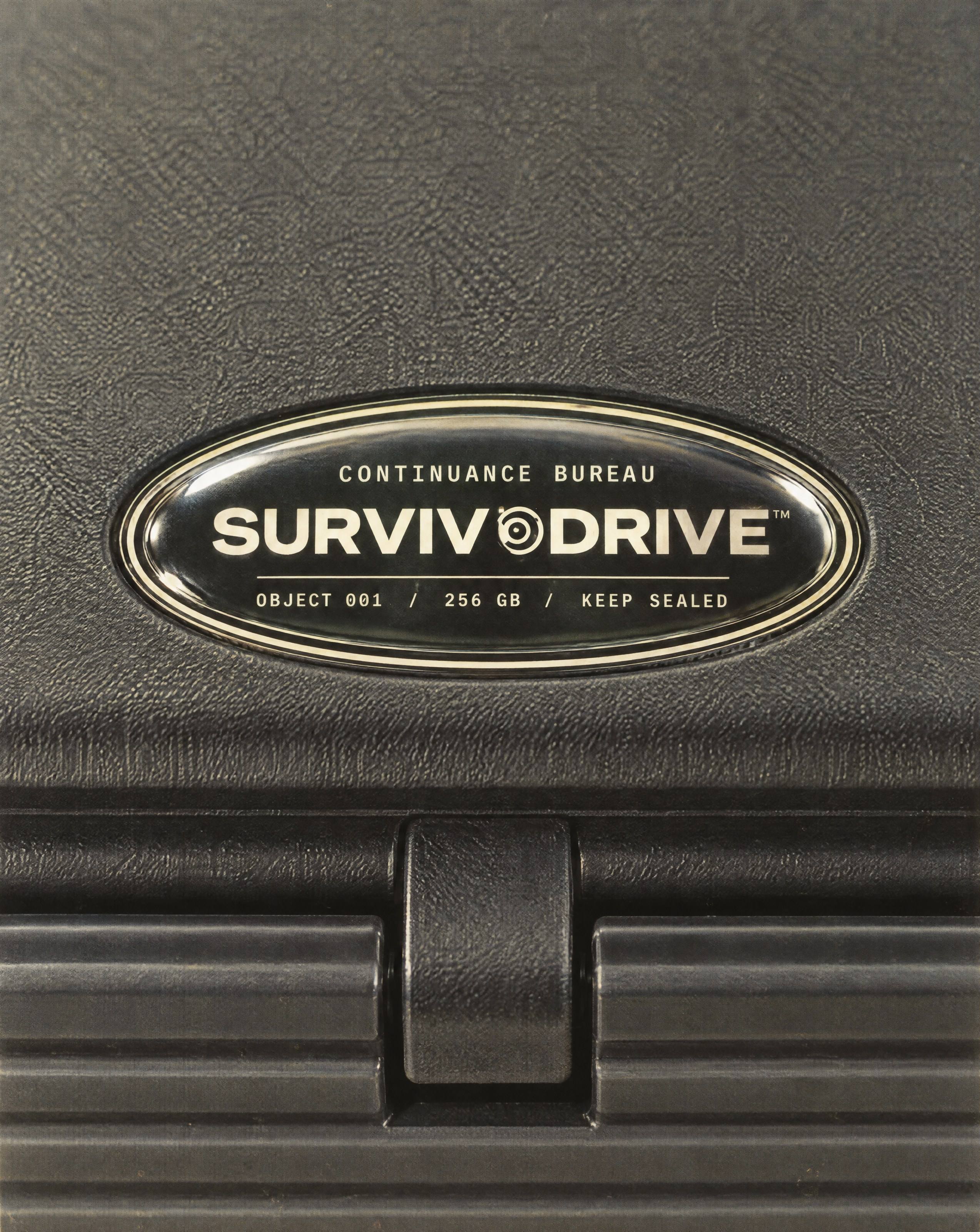 Macro of the domed resin SURVIVODRIVE badge in the oval indent on the front of the case lid, just behind the latch